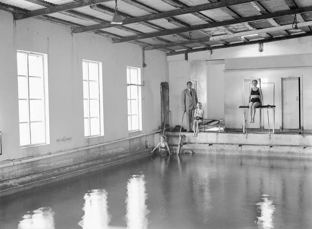 Interior despido de uma piscina coberta com teto de chapa: um homem de fato e gravata segura uma vara comprida na borda, uma criança está na água junto à escada, duas raparigas sentadas no trampolim
