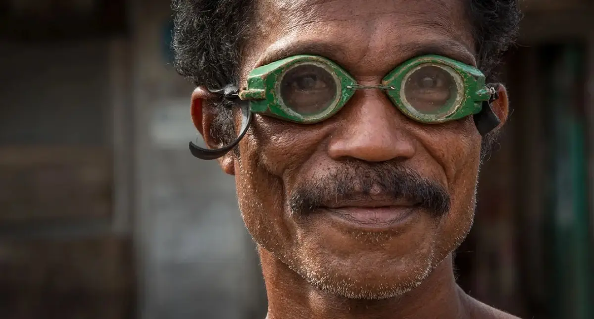 Primeiro plano de um pescador Bajau que usa óculos esculpidos à mão na madeira e pintados de verde, com duas lentes de vidro redondas mantidas por um fio metálico e uma fita de borracha atrás das orelhas