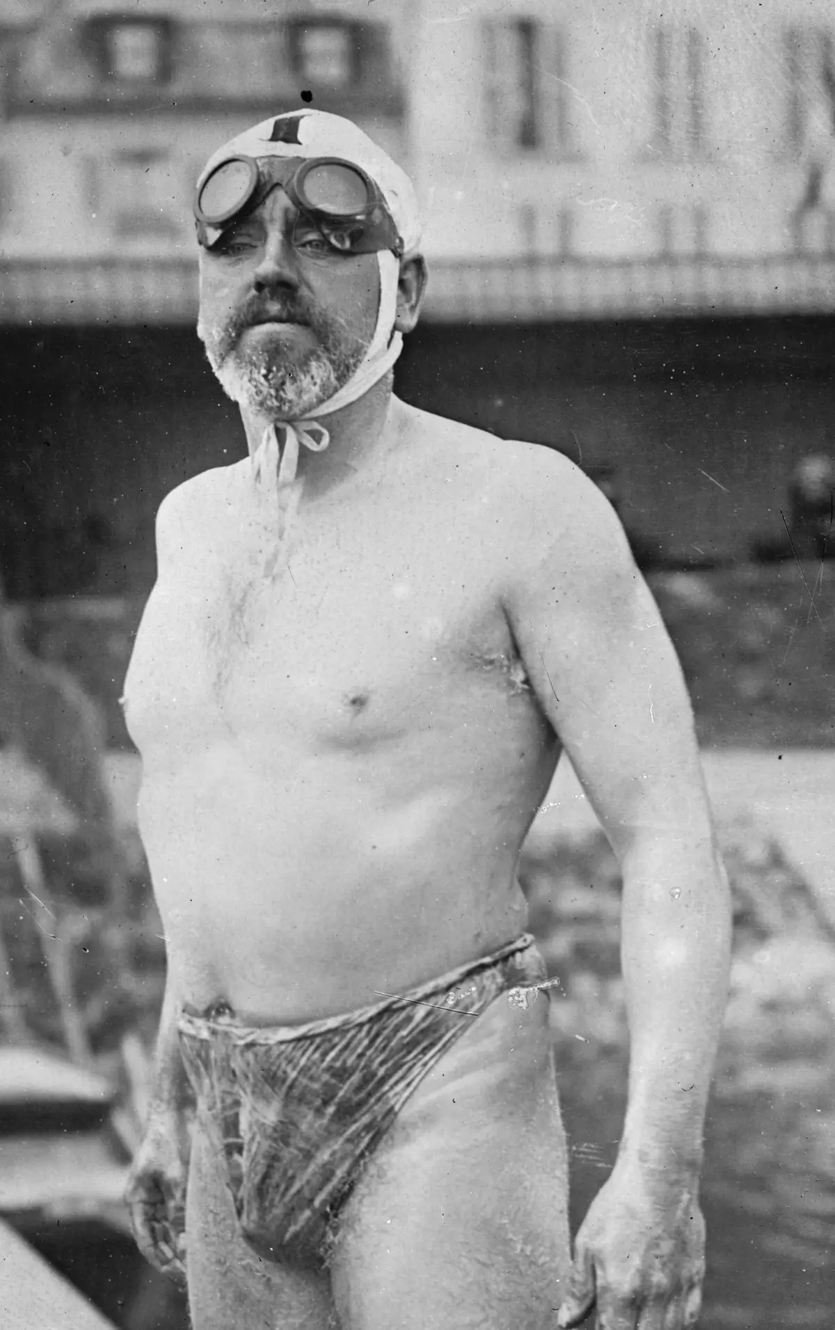Bill Burgess de pé num pontão em 1911, o corpo coberto de gordura, o fato de banho de lã e os óculos de motociclista levantados na testa sobre a touca atada debaixo do queixo