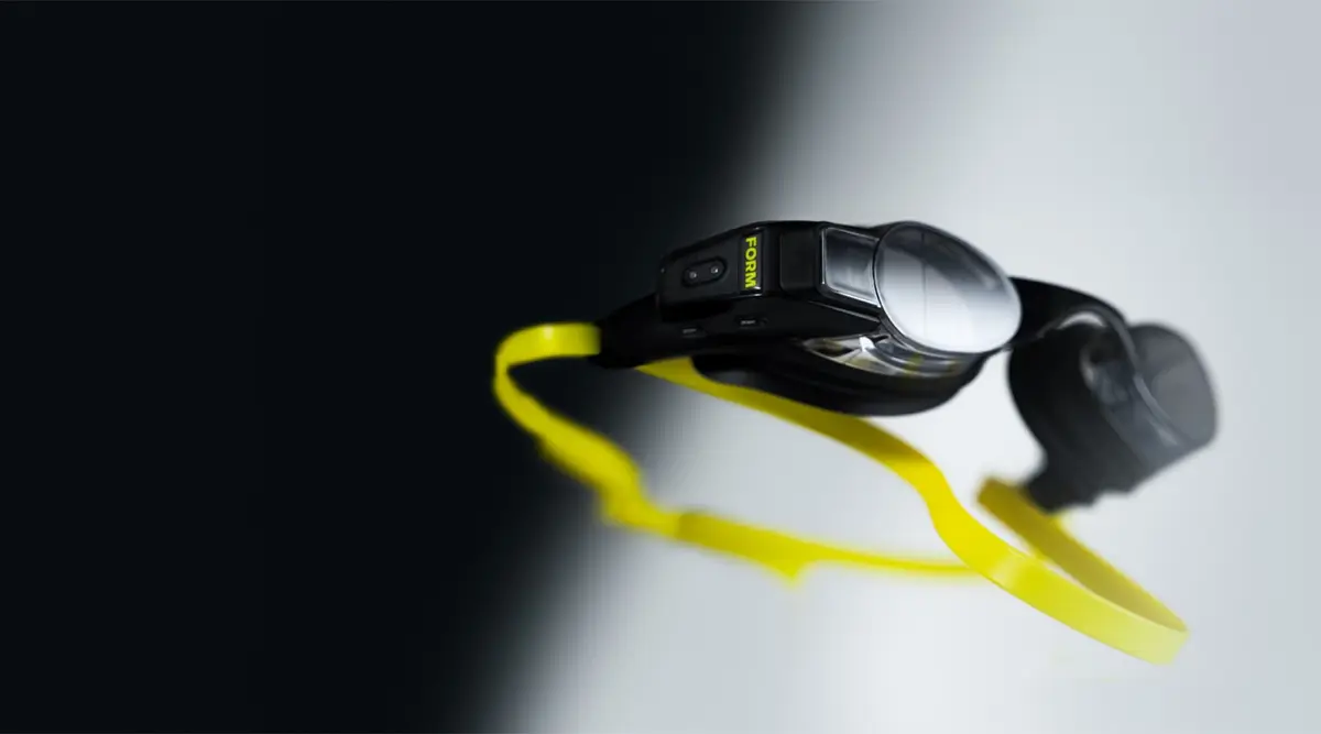 Um par de óculos FORM Smart Swim sobre fundo preto e branco: o módulo da eletrónica sobressai do lado da lente direita, a fita é amarela