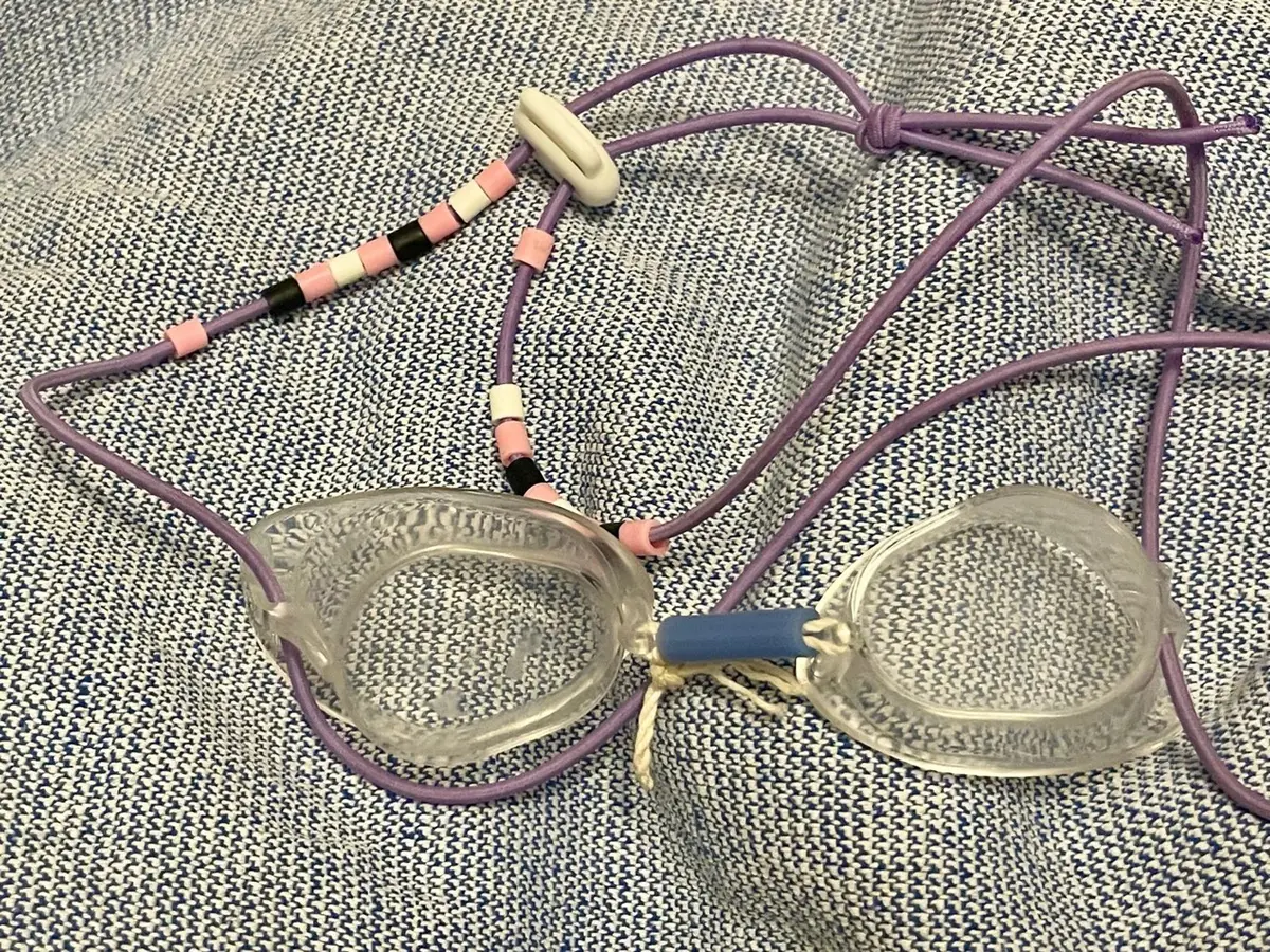 Um par de óculos suecos montados à mão sobre uma toalha: duas lentes transparentes nuas, o cordão roxo com os espaçadores enfiados um a um, um tubinho de borracha e um fio atado no lugar da ponte nasal