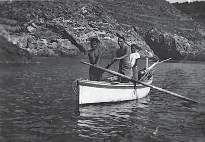 Fotografía en blanco y negro: tres hombres de pie sobre un gozzo blanco amarrado bajo una costa rocosa, con las largas cañas de bambú apoyadas de lado en la barca