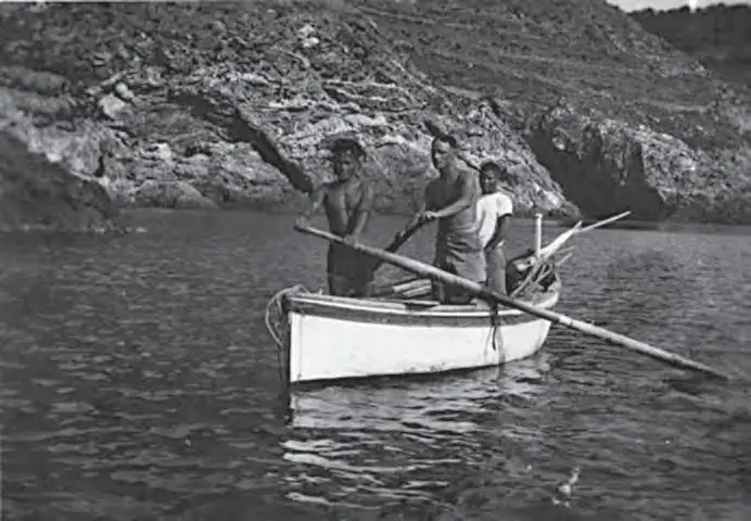 Fotografia a preto e branco: três homens de pé num barco de pesca branco ancorado sob uma costa rochosa, com as longas canas de bambu apoiadas transversalmente no barco