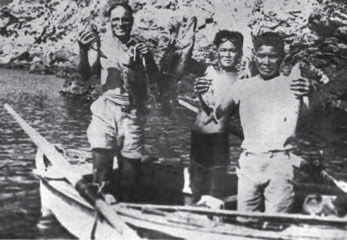 Fotografía en blanco y negro: tres hombres de pie sobre una barca levantan meros recién pescados; el del centro lleva las gafas de madera subidas sobre la frente
