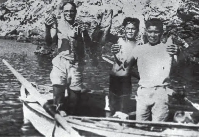 Fotografia a preto e branco: três homens de pé num barco levantam garoupas recém-pescadas; o do centro traz os óculos de madeira levantados na testa