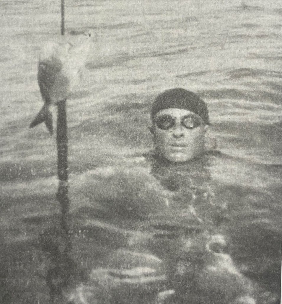 Fotografía en blanco y negro: un hombre con gorro oscuro y gafas redondas emerge del agua junto a una lanza con un pez ensartado
