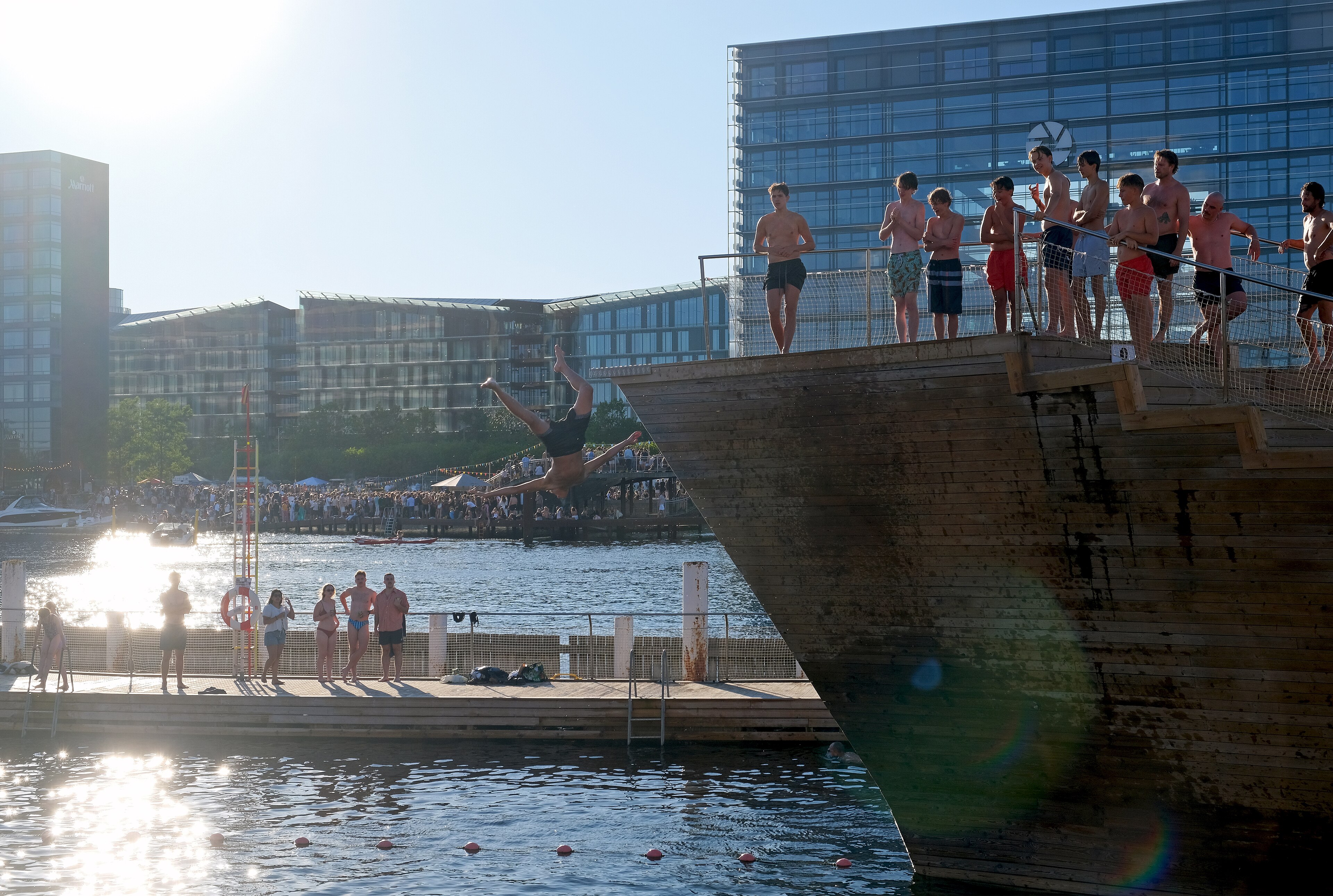 Un grupo de personas se zambulle desde el muelle del baño portuario de Islands Brygge en Copenhague, con el agua verde oscuro del canal y la ciudad de fondo